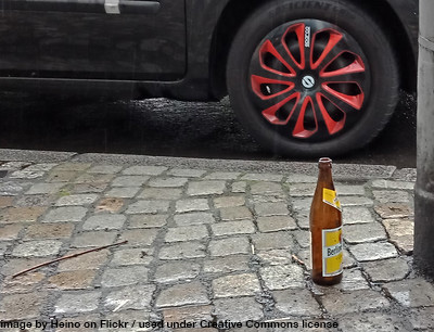 A beer bottle on a cobbled sidewalk near the wheel of a car