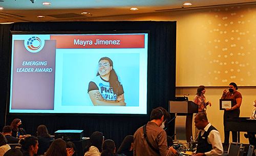 a large screen shows a picture of Mayra Jimenez with the caption "emerging leader award," while besides it Mayra stands with another woman, receiving a plaque